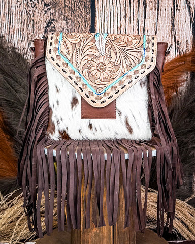 The Straybird Hair on Hide Fringe Crossbody Bag displayed on a wooden table with wheat behind it, sitting in front of a wood back drop.