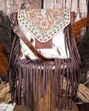 The Straybird Hair on Hide Fringe Crossbody Bag displayed on a wooden table with wheat behind it, sitting in front of a wood back drop.