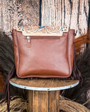 The Straybird Hair on Hide Fringe Crossbody Bag, turned around to show the back of the purse which is made of leather. There is a zipper on the top. It is being displayed on a wooden table with wheat behind it, sitting in front of a wood back drop.