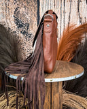 Side view of the Straybird Hair on Hide Fringe Crossbody Bag, displayed on a wooden table with wheat behind it. It is sitting in front of a wood back drop.