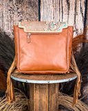 The Sunny Jo Fringe Crossbody Bag, turned around to show the leather back. There is a zipper on the top. Wheat and a wood back drop are behind it.