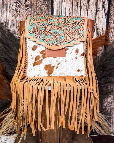 The Sierra Azul Hair on Hide Fringe Crossbody Bag displayed on a wooden table with wheat behind it, sitting in front of a wood back drop.