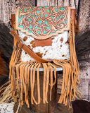 The Sierra Azul Hair on Hide Fringe Crossbody Bag displayed on a wooden table with wheat behind it, sitting in front of a wood back drop.