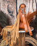 Side view of the Sierra Azul Hair on Hide Fringe Crossbody Bag, displayed on a wooden table with wheat behind it. Sitting in front of a wood back drop.