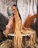 Side view of the Sierra Azul Hair on Hide Fringe Crossbody Bag displayed on a wooden table with wheat behind it. It is sitting in front of a wood back drop.