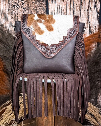 The Mae Hair on Hide Fringe Crossbody Bag displayed on a wooden table with wheat behind it, sitting in front of a wood back drop.