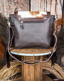 The Mae Hair on Hide Fringe Crossbody Bag, turned around to show the leather back. There is a zipper on the top. It is being displayed on a wooden table with wheat and a wood back drop behind it.