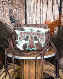 The Quill Hair on Hide Fringe Crossbody Bag displayed on a wooden table with wheat behind it, sitting in front of a wood back drop.