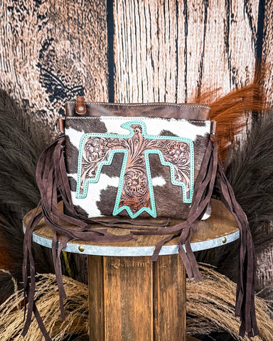 The Quill Hair on Hide Fringe Crossbody Bag displayed on a wooden table with wheat behind it, sitting in front of a wood back drop.