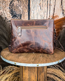 The Quill Hair on Hide Fringe Crossbody Bag turned around to show the leather back. There is a zipper on the top. It is being displayed on a wooden table with wheat and a wood back drop behind it.