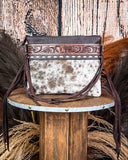 The Ardelle Hair on Hide Fringe Crossbody Bag displayed on a wooden table with wheat behind it, sitting in front of a wood back drop.