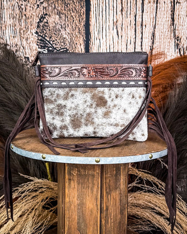 The Ardelle Hair on Hide Fringe Crossbody Bag displayed on a wooden table with wheat behind it, sitting in front of a wood back drop.