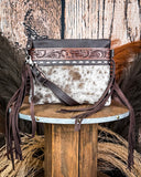 The Ardelle Hair on Hide Fringe Crossbody Bag displayed on a wooden table with wheat behind it, sitting in front of a wood back drop.
