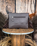 The Ardelle Hair on Hide Fringe Crossbody Bag, turned around to show the back of the purse which is made of leather. There is a zipper on the top. There's a wood backdrop and wheat grass behind it.