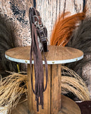 Side view of the Ardelle Hair on Hide Fringe Crossbody Bag, being displayed on a wooden table. The fringe on the bag is cascading off the side of the table. There's a wood back drop and wheat grass behind it.