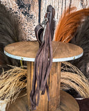 Side view of the Ardelle Hair on Hide Fringe Crossbody Bag, being displayed on a wooden table. The fringe on the bag is cascading off the side of the table. There's a wood back drop and wheat grass behind it.