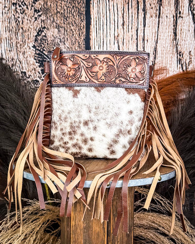 The Lariat Hair on Hide Fringe Crossbody Bag displayed on a wooden table with wheat grass and a wood backdrop behind it.
