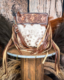 The Lariat Hair on Hide Fringe Crossbody Bag displayed on a wooden table with wheat grass and a wood back drop behind it.