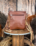 The Lariat Hair on Hide Fringe Crossbody Bag, turned around to show the back of the bag, which is made of leather. The bag is being displayed on a wooden table with wheat grass and a wood backdrop behind it.