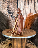 Side view of the Lariat Hair on Hide Fringe Crossbody Bag, displayed on a wooden table with wheat grass and a wood back drop behind it.