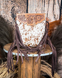 The Roux Hair on Hide Fringe Crossbody Bag, displayed on a wooden table with wheat grass and a wood backdrop behind it.