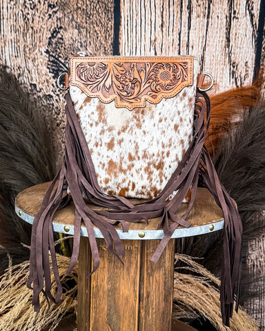 The Roux Hair on Hide Fringe Crossbody Bag, displayed on a wooden table with wheat grass and a wood backdrop behind it.