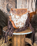 The Roux Hair on Hide Fringe Crossbody Bag, displayed on a wooden table with wheat grass and a wood backdrop behind it.