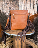 The Roux Hair on Hide Fringe Crossbody Bag, turned around to show the back of the bag, which is made of leather. There is a zipper at the top. It is being displayed on a wooden table with wheat grass and a wood back drop behind it.
