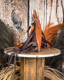 Side view of the Roux Hair on Hide Fringe Crossbody Bag, being displayed on a wooden table with wheat grass and a wood backdrop behind it.