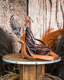 Side view of the Roux Hair on Hide Fringe Crossbody Bag, being displayed on a wooden table with wheat grass and a wood back drop behind it.