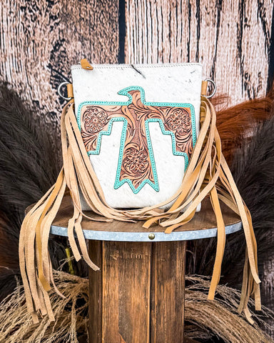 The Talon Hair on Hide Fringe Crossbody Bag, displayed on a wooden table with wheat grass and a wood backdrop behind it.