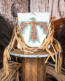 The Talon Hair on Hide Fringe Crossbody Bag, displayed on a wooden table with wheat grass and a wood backdrop behind it.