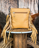 The Talon Hair on Hide Fringe Crossbody Bag, turned around to show the back of the bag, which is made of leather. There is a zipper at the top. It is being displayed on a wooden table with wheat grass and a wood back drop behind it.