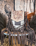 The Reveena Hair on Hide Fringe Phone Crossbody Bag, displayed on a wooden table with wheat grass and a wood back drop behind it.