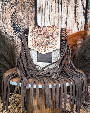The Reveena Hair on Hide Fringe Phone Crossbody Bag, displayed on a wooden table with wheat grass and a wood back drop behind it.