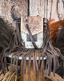 The Reveena Hair on Hide Fringe Phone Crossbody Bag, displayed on a wooden table with wheat grass and a wood backdrop.