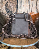 TheReveena Hair on Hide Fringe Phone Crossbody Bag, turned around to show the back of the bag, which is made of leather. There is a zipper at the top. Wheat grass and a wood backdrop is behind it.