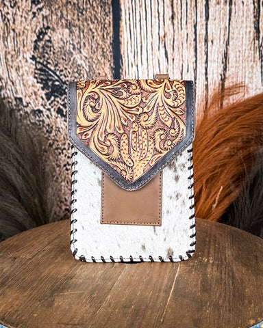The Vesper Hair on Hide Phone Crossbody Bag, displayed on a wooden table with wheat grass and a wood backdrop behind it.