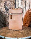 The Vesper Hair on Hide Phone Crossbody Bag, turned around to show the back of the bag which is made of leather. There is a zipper at the top. It is being displayed on a wooden table. There is wheat grass and a wood backdrop behind it.