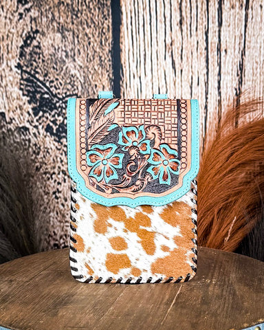 The Lark Hair on Hide Phone Crossbody Bag, displayed on a wooden table with wheat grass and a wood backdrop behind it.