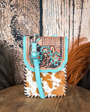 The Lark Hair on Hide Phone Crossbody Bag, displayed on a wooden table with wheat grass and a wood backdrop behind it.