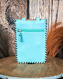 The Lark Hair on Hide Phone Crossbody Bag, turned around to show the back of the bag, which is made of leather. There is a zipper on the top. It is being displayed on a wooden table with wheat grass and a wood backdrop behind it.