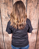 A girl wearing the Four Aces Graphic Tee, facing the wall behind her to show the back of the Four Aces Graphic Tee which is blank.