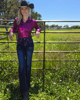 A woman wearing a shiny magenta western-style button-up shirt, dark blue jeans, a black cowboy hat, and boots stands outdoors in a grassy field with a metal fence and trees in the background.
