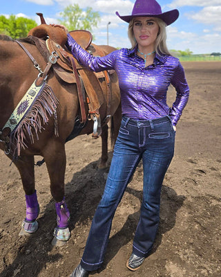 A woman wearing a shiny purple long-sleeve button-up shirt, blue jeans, and a purple cowboy hat stands beside a brown horse in an outdoor dirt arena.