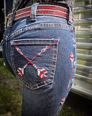 Close-up of the back of blue denim jeans worn by a person, featuring red and white embroidered crossed arrows on the back pocket and a matching red-and-white embroidered belt.
