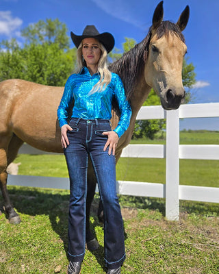 A woman wearing a shiny metallic blue long-sleeve shirt, dark blue jeans, and a black cowboy hat stands outdoors next to a brown horse in front of a white fence.