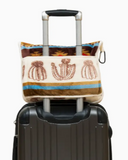 the bagged Cactus Stripe Travel Blanket hooked onto a suitcase by its opening in a white backdrop.