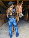 Woman in cowboy hat and blue jeans standing next to a brown horse in a stable.