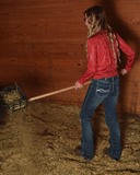 Person in a red jacket and blue jeans using a hay fork in a barn setting.